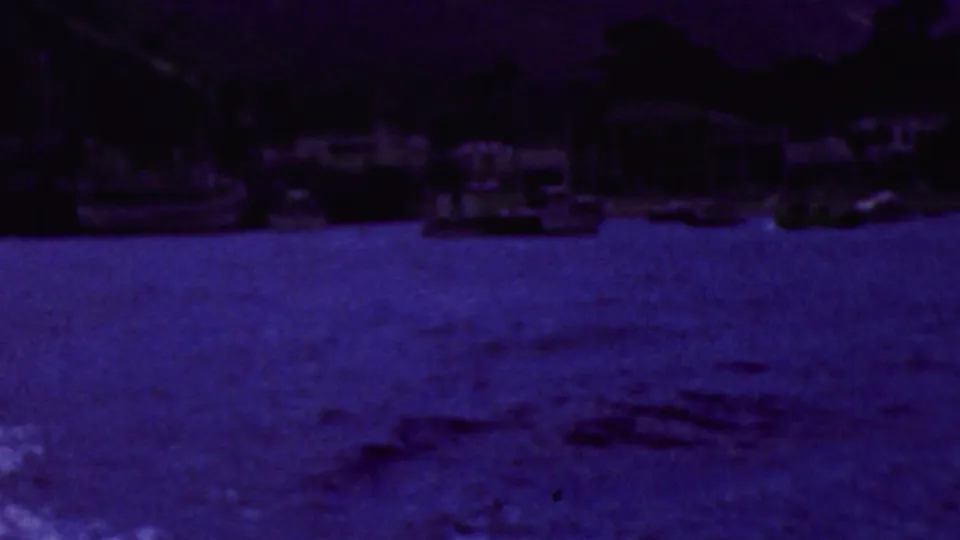1961 California home-movie frame showing rippling water from a boat with a distant shoreline, houses, and moored small boats under heavy purple-blue color shift.