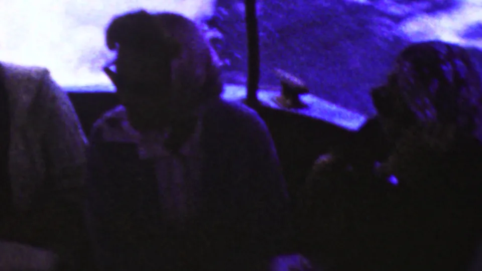 Low-light 8mm frame of passengers seated aboard the boat, including a person in dark glasses and headscarved figures, with bright water visible through the window openings.