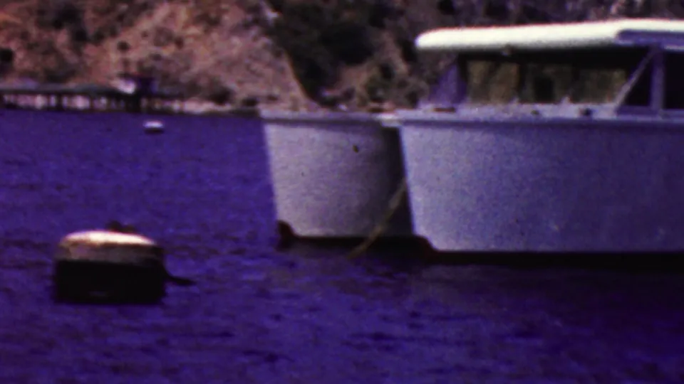 A white boat sits on calm water near a rocky shoreline, providing dockside or waterfront context for the 1961 California home-movie clip.