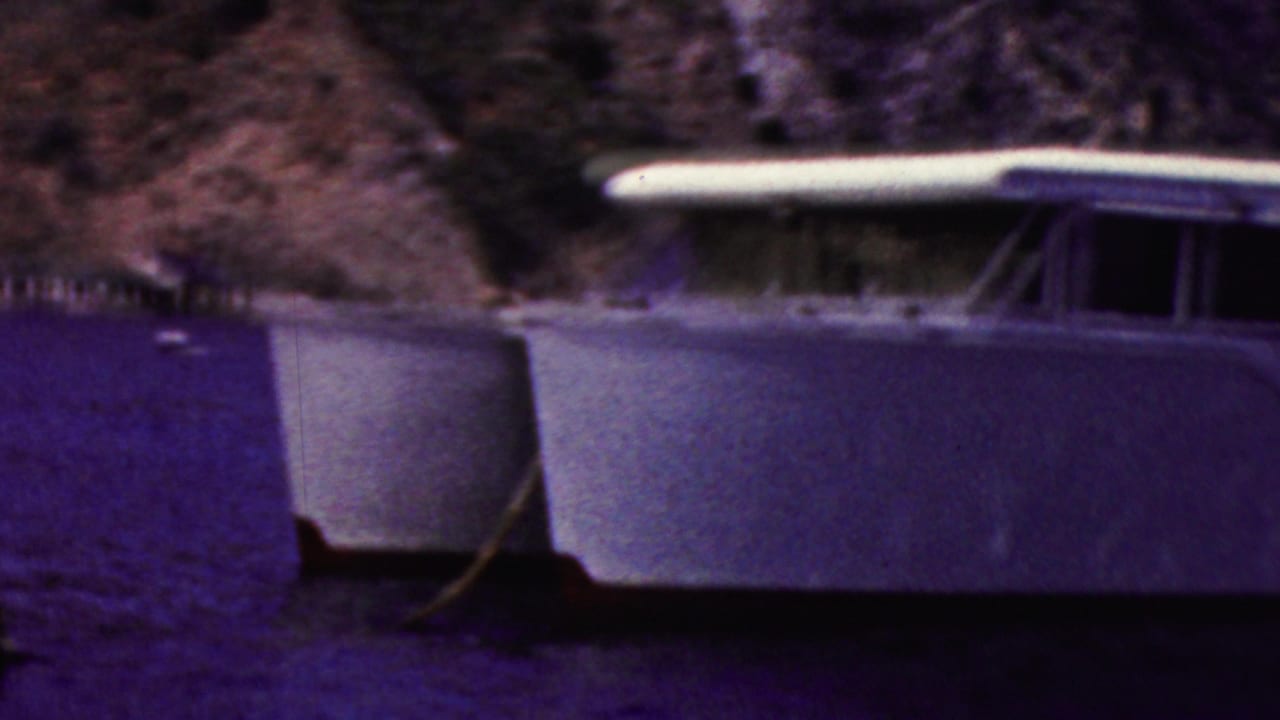 1961 California 8mm frame showing the bow and cabin side of a white motorboat at anchor on dark coastal water, with rocks behind it.