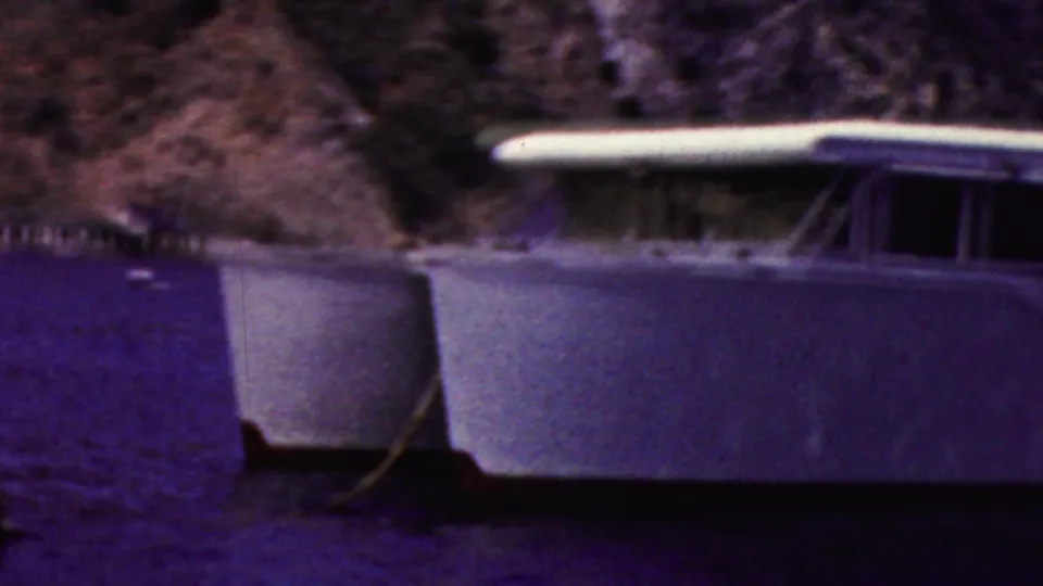 1961 California 8mm frame showing the bow and cabin side of a white motorboat at anchor on dark coastal water, with rocks behind it.