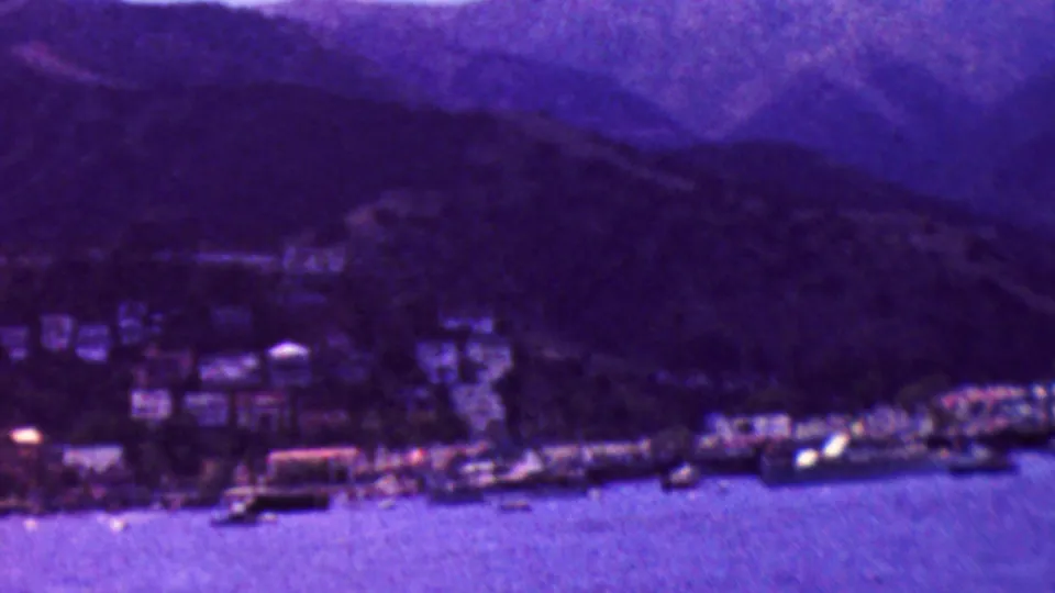 1961 California 8mm travel view of a lakeside town, docked boats, shoreline buildings, water, and mountain ridges under a strong purple color shift.