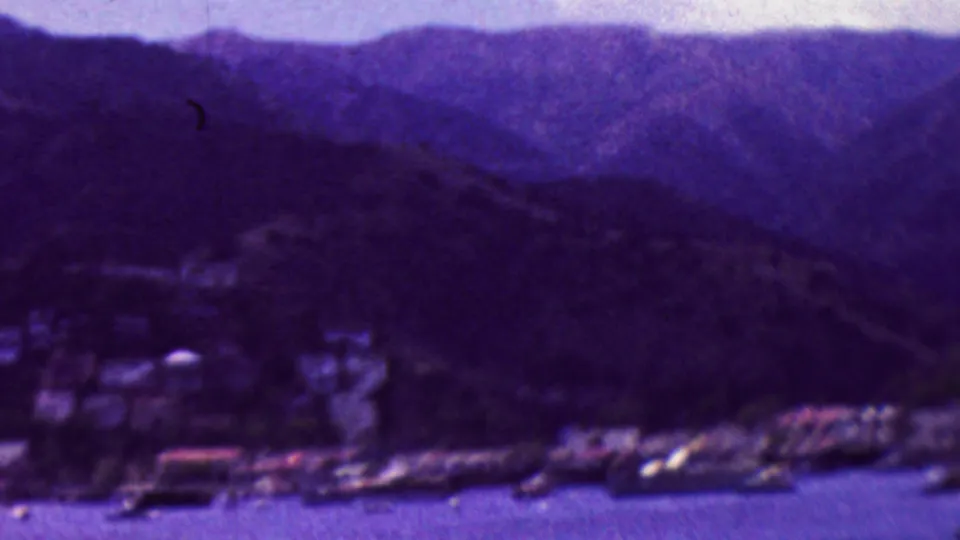 Soft 8mm frame of a California waterfront town with boats near the shore, clustered buildings, and layered mountain ridges in the background.