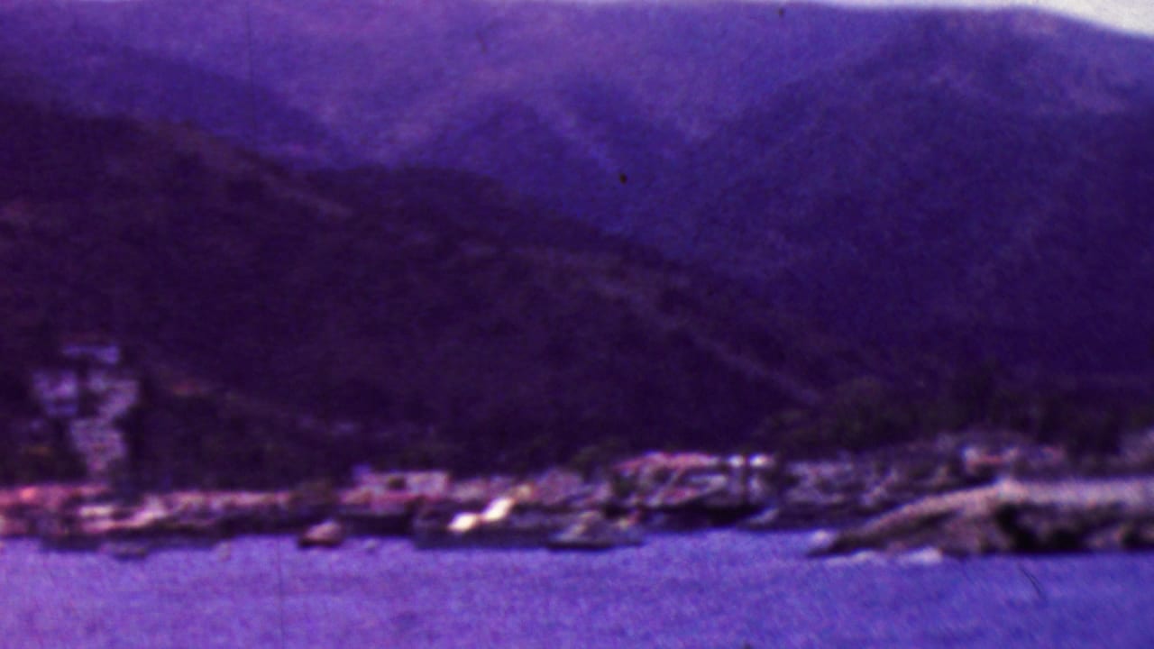 1961 California 8mm frame showing a soft purple coastal view with water in the foreground, small shoreline buildings or boats, and dark mountain ridges in the background.
