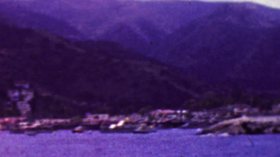 1961 California 8mm frame showing a soft purple coastal view with water in the foreground, small shoreline buildings or boats, and dark mountain ridges in the background.