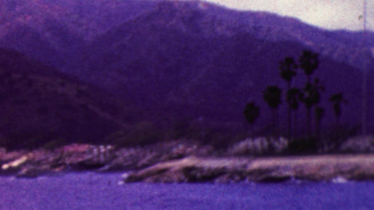 1961 California 8mm frame of a waterfront community with palm trees near the shore, boats along the water, and mountains rising behind the coast.