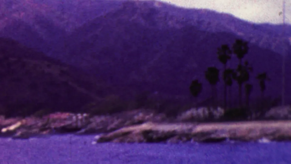 1961 California 8mm frame of a waterfront community with palm trees near the shore, boats along the water, and mountains rising behind the coast.