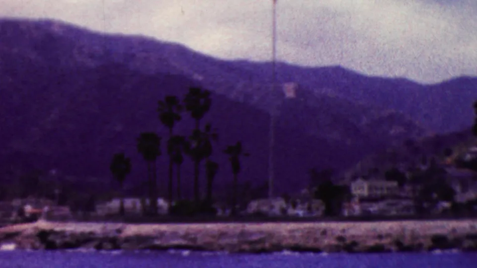 1961 California 8mm frame showing a palm-lined shoreline with small waterfront structures, boats near the edge of the water, and mountain slopes in the background.