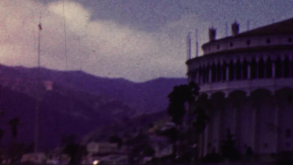 1961 California 8mm frame with a rounded multi-story waterfront building at the right edge, palm trees beside it, and mountain ridges across the background.