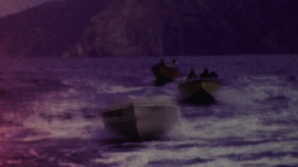 Several small motorboats with distant passengers move across rough coastal water near a dark rocky cliff, with a soft purple cast from the aged 8mm film.