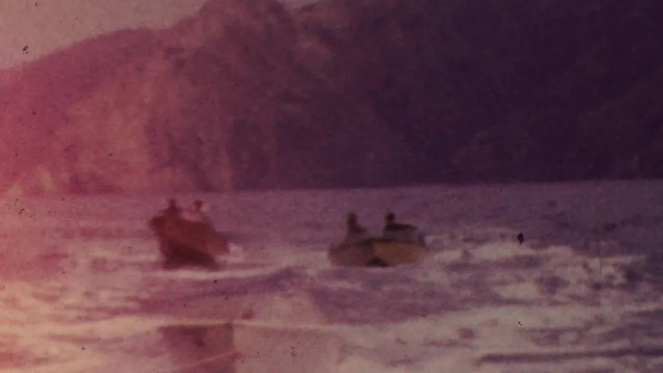 Two small motorboats with distant riders cross uneven ocean water below a rocky California coastline, with pink color fading and visible film grain.