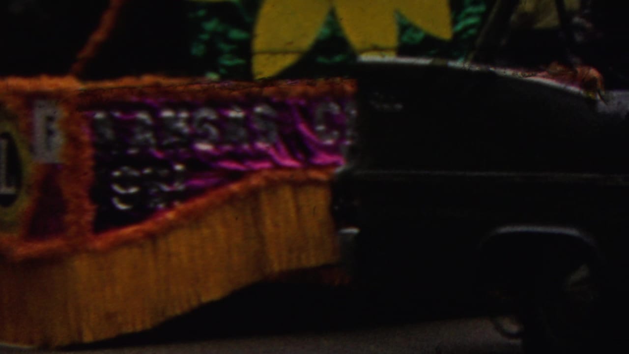 1961 California home-movie frame showing a decorated parade float with metallic purple trim, gold fringe, partial lettering, and a dark vehicle passing in front.