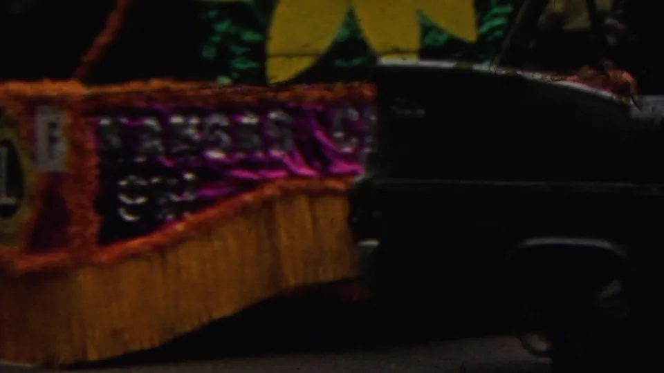 1961 California home-movie frame showing a decorated parade float with metallic purple trim, gold fringe, partial lettering, and a dark vehicle passing in front.