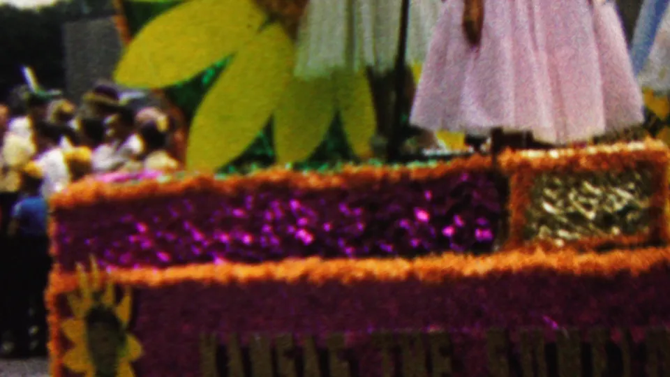 1961 California parade float frame with sunflower cutouts, purple and gold trim, seated riders, and a woman in a light pink dress standing near the float edge.