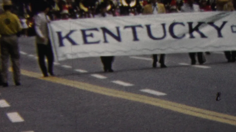 1961 California parade frame showing a large Kentucky banner beside uniformed marching band members, with spectators and brass instruments partly visible behind it.