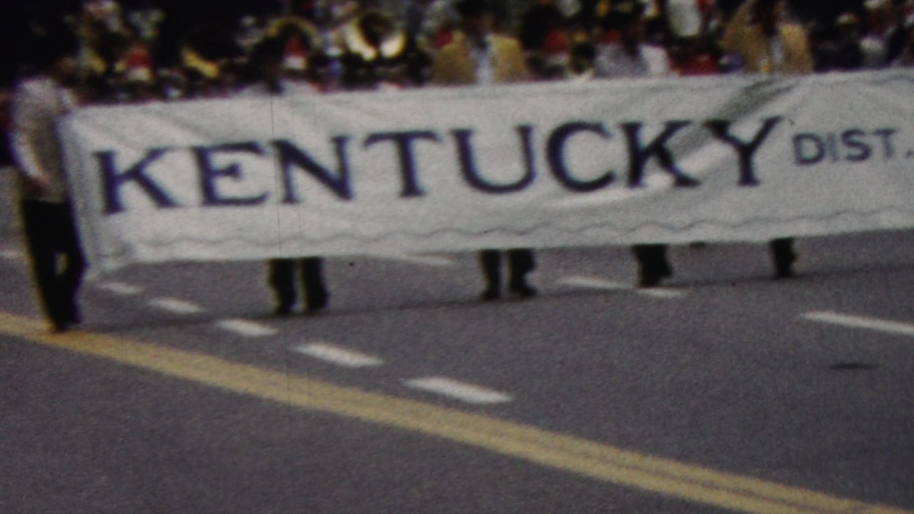 In 1961 California parade footage, marchers carry a large white banner reading Kentucky Dist. 4.3 Lions while spectators stand behind them.