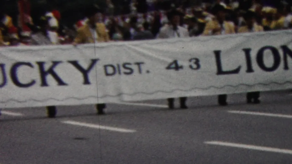 The home-movie frame catches the Kentucky Lions banner mid-passage, with District 4.3 text, road lines, and legs of marchers visible below.