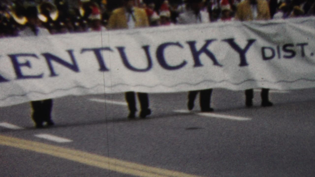 In California 1961 footage, marchers carry a large cloth banner reading Kentucky district while spectators and other parade participants fill the street behind them.