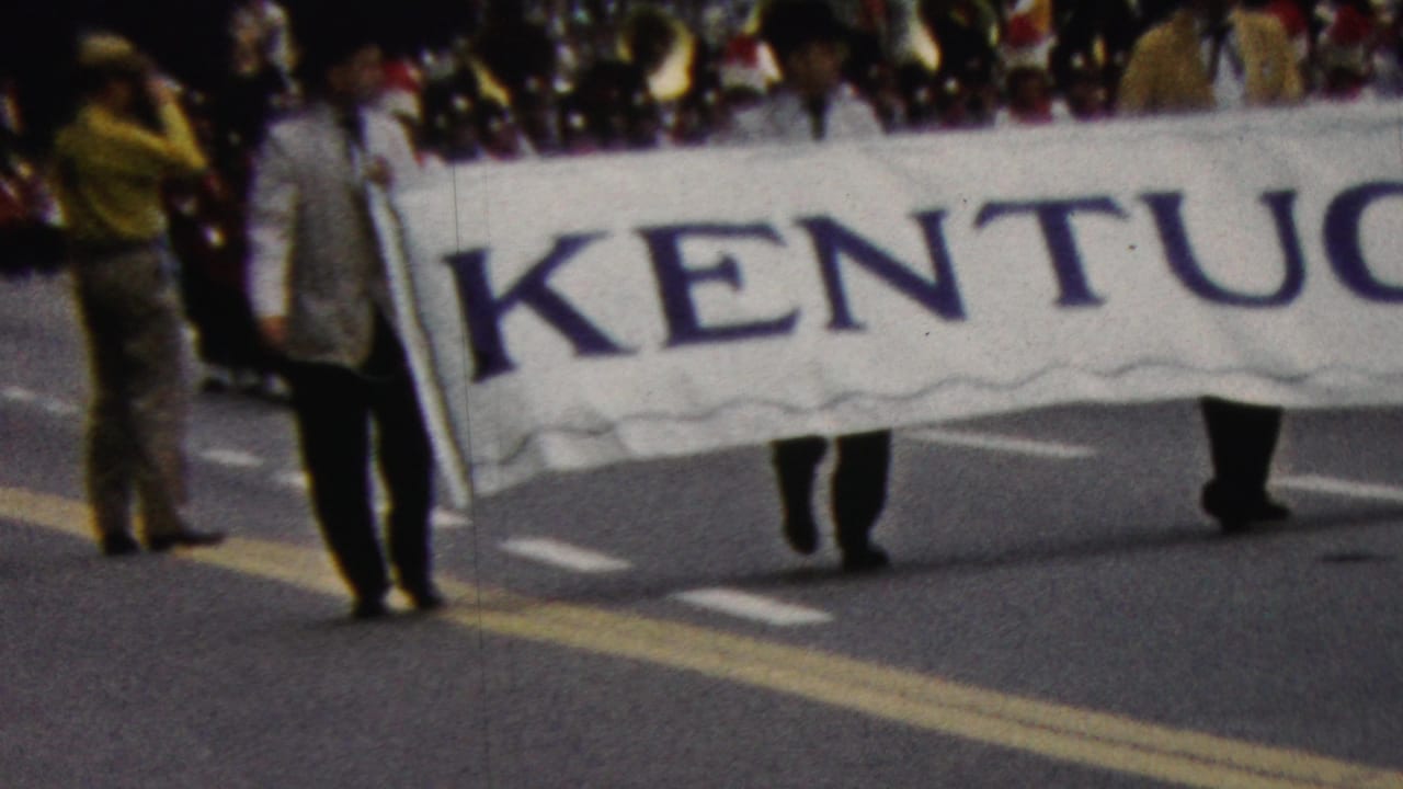 A Kentucky banner continues across the street with marchers and spectators behind it.