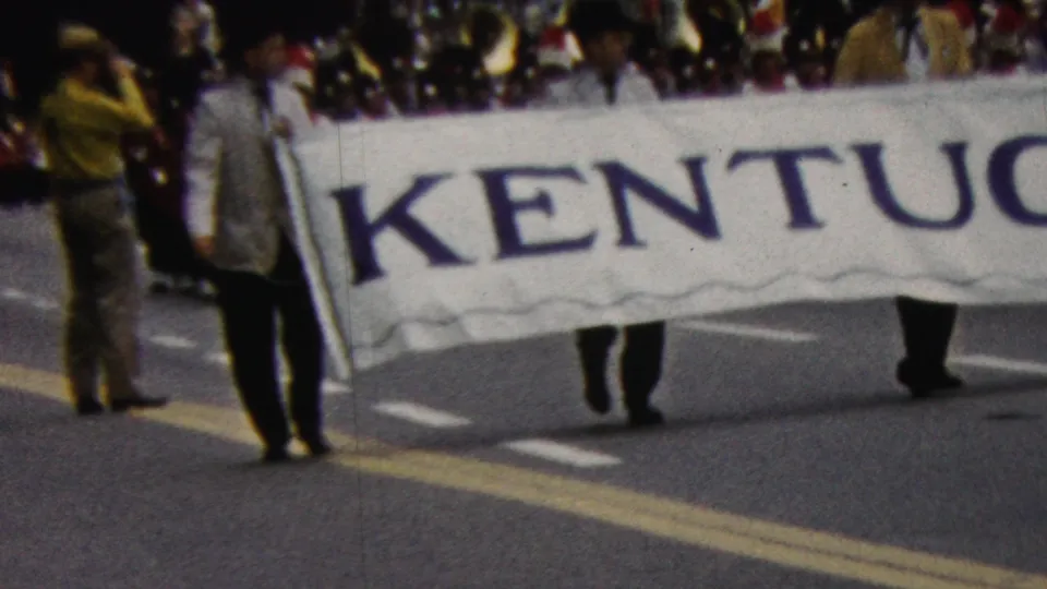 The Kentucky-labeled parade banner moves past the camera as marchers hold it at street level and a dense crowd stands along the route in the background.