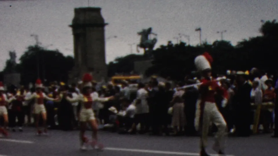 In the 1961 California parade scene, red-and-white uniformed majorettes and band members move along the street before spectators, with a stone tower and equestrian statue visible in the background.