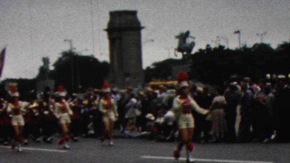 The closing frame shows marching performers in red-and-white uniforms moving past a large spectator crowd, with a civic tower and equestrian statue behind the parade route.