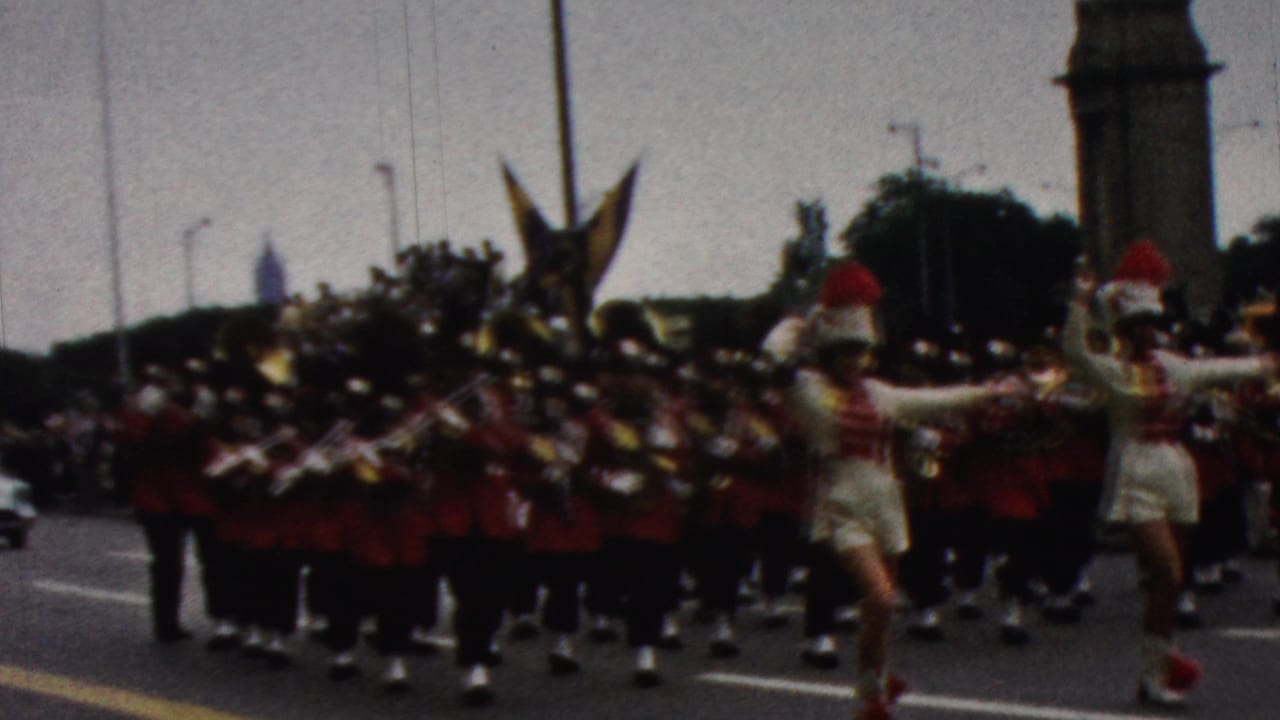 1961 California home-movie frame showing cheerleaders leading a red-uniformed marching band down a city street with spectators and a tall monument in the distance.