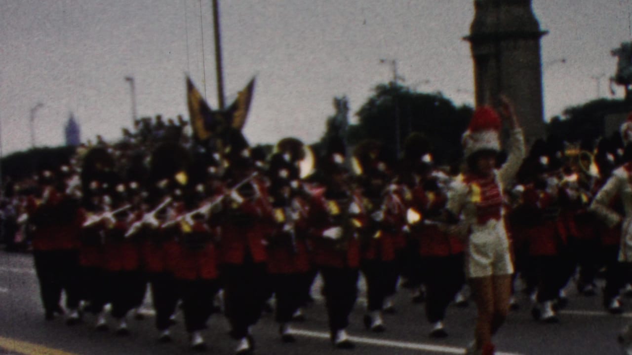 Cheerleaders and a red-uniformed marching band pass the camera in parade formation.
