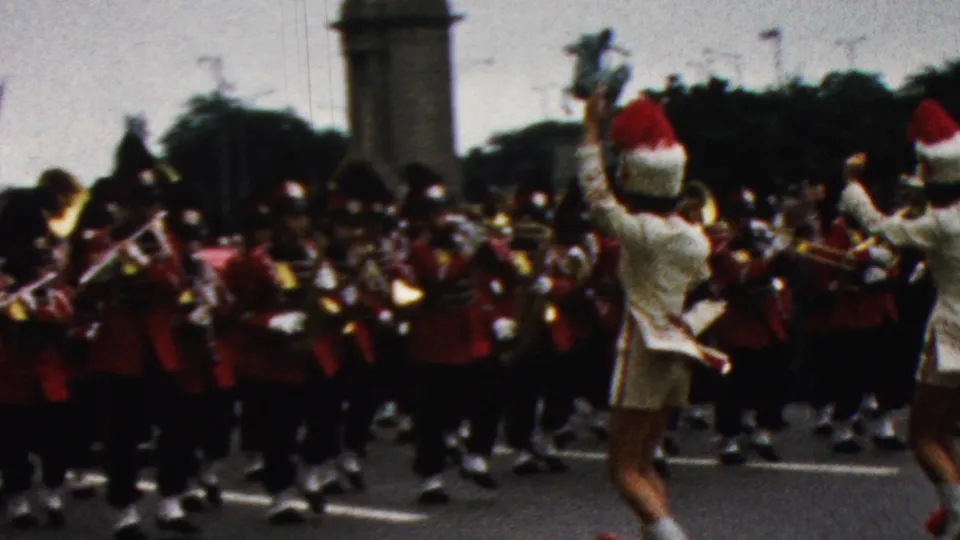 From a spectator viewpoint, two cheerleaders in white uniforms face a red-uniformed marching band during a 1961 California street parade.