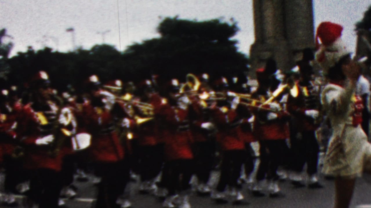 1961 California home-movie frame showing a red-uniformed marching band with brass instruments passing a blue decorated float at a parade.