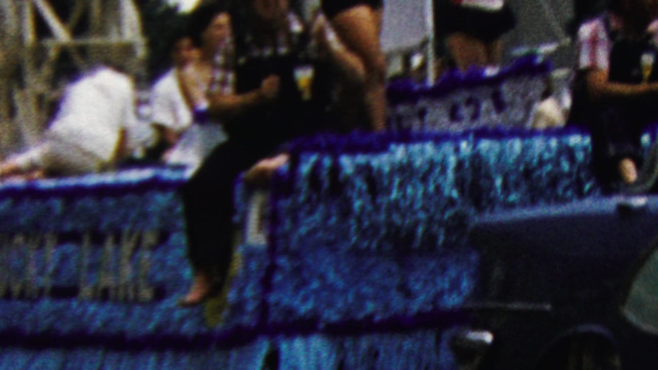 Blue parade float with riders and nearby baton twirler beside a marching band.