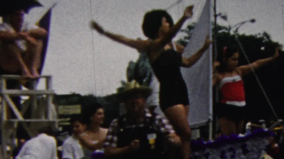 1961 California home-movie frame showing a majorette raising a baton near spectators, parade rigging, and street-side background elements.