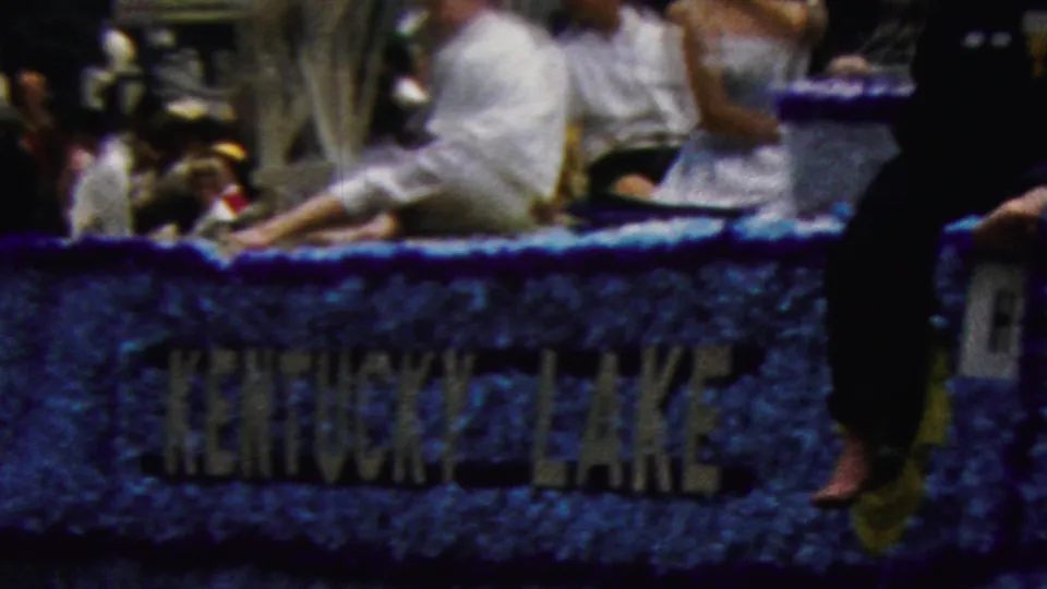 1961 California parade frame showing a blue tinsel-covered float with readable Kentucky Lake lettering and riders seated along the edge.