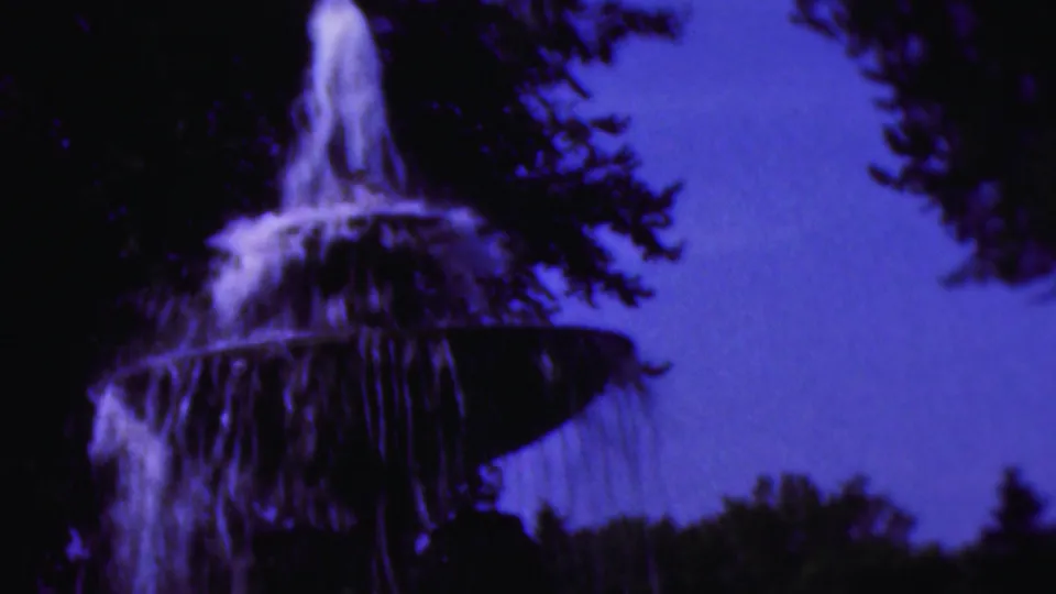 1954 Wisconsin 8mm frame showing the top of a tiered outdoor fountain with water spilling over dark bowls beneath silhouetted trees and a blue-purple sky.