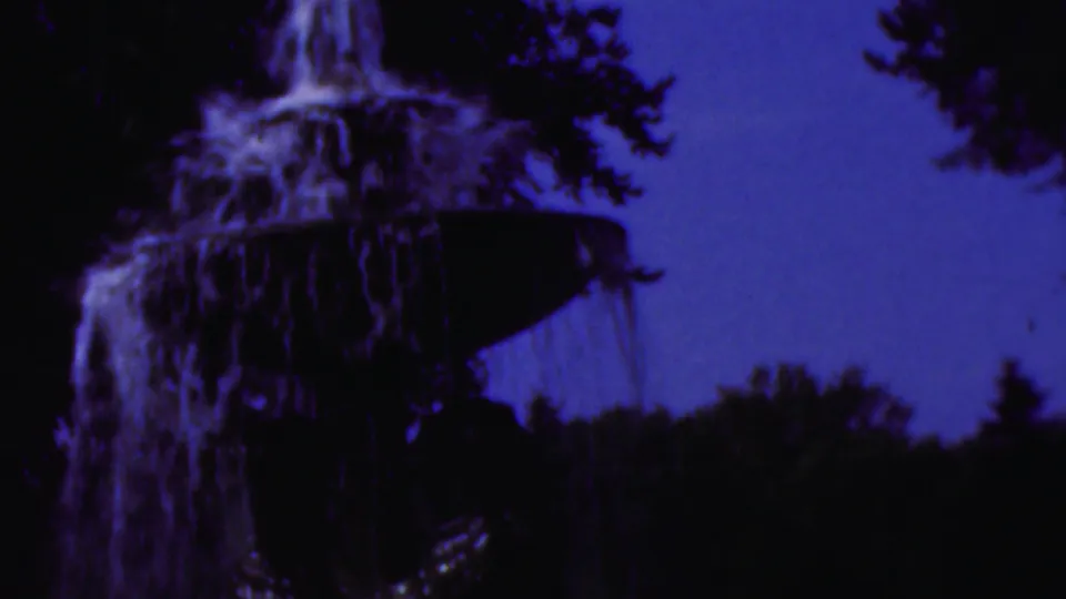 1954 Wisconsin 8mm frame with a closer view of the fountain's upper bowl, cascading water, dark surrounding trees, and strong purple-blue film color cast.