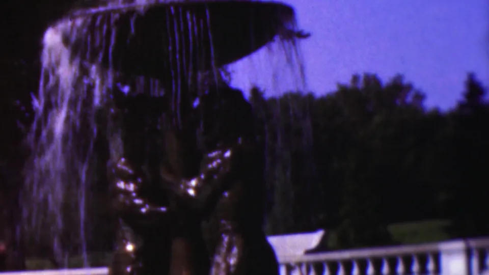 1954 Wisconsin 8mm frame tilted down to show falling fountain water, a dark reflective sculptural base, lawn, trees, and a pale balustrade or railing.