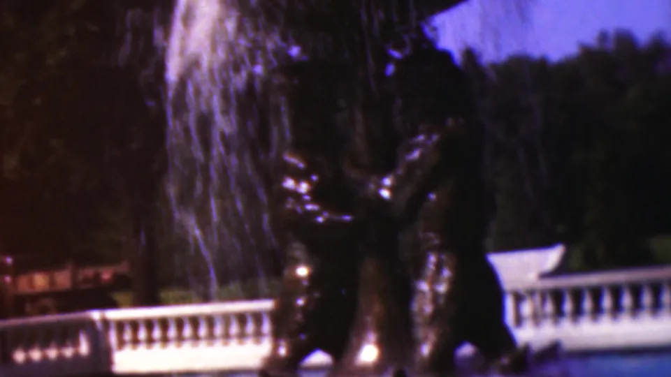 1954 Wisconsin 8mm frame showing the lower fountain, falling water, dark reflective sculptural form, tree line, lawn, and pale balustrade in soft purple-blue color.