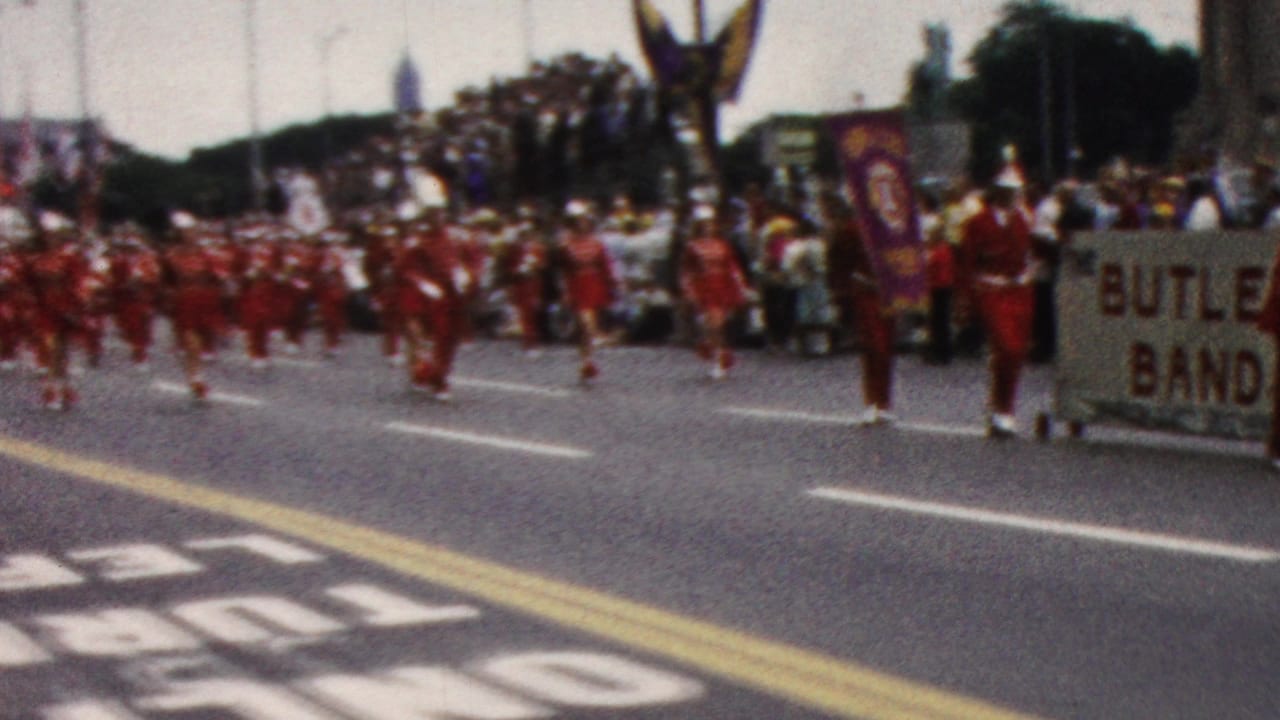1961 California home-movie frame showing red-uniformed members of the Butler Band approaching along a street parade route as spectators watch from the sidewalk.