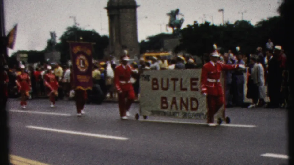 1961 California home-movie frame showing a wheeled Butler Band banner, a purple Lions Club flag, red-uniformed parade members, and spectators near the curb.