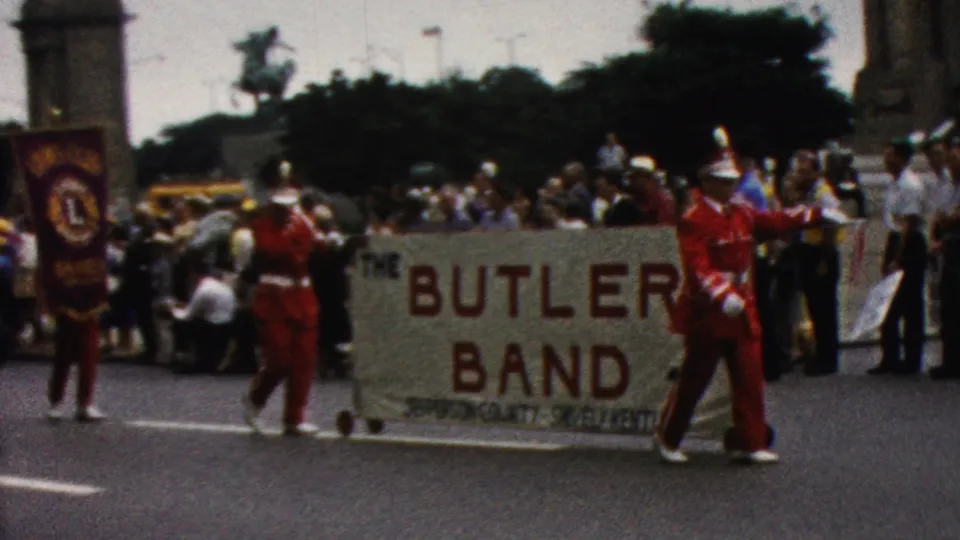 1961 California frame with the Butler Band banner prominent in the foreground, red-uniformed marchers nearby, and spectators watching from the sidewalk behind.