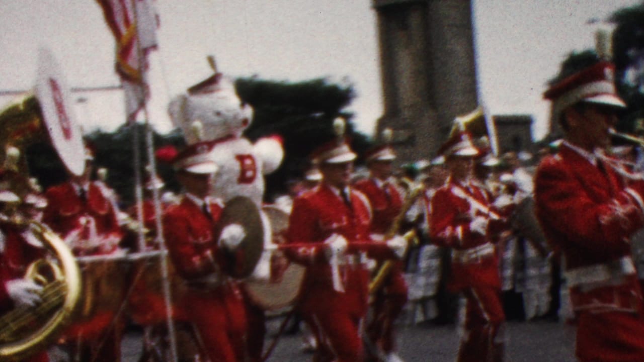 1961 California 8mm frame showing a red-uniformed marching band with white plumed hats, drums, brass instruments, flags, and spectators along a city street.
