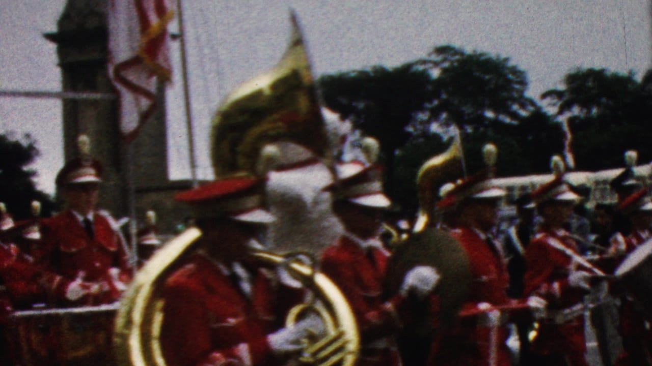 Band members in red uniforms march past with tubas, drums, and a large white mascot figure nearby.