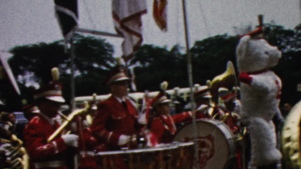 1961 California 8mm frame showing percussion and brass players in red uniforms beside a large white mascot figure, with flags and spectators visible in the parade background.