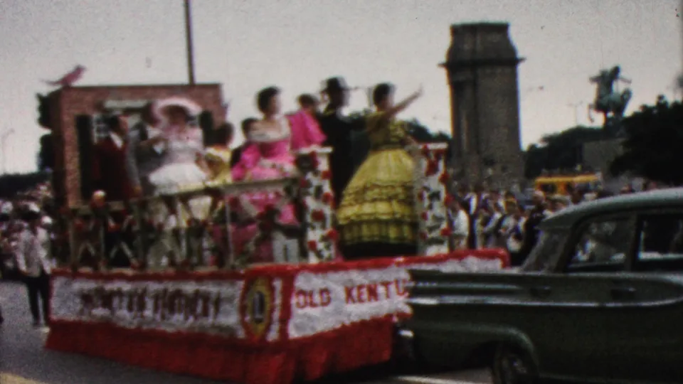 1961 California parade frame showing a red-and-white decorated float with costumed riders, partial lettering on the float side, spectators, civic architecture, and a dark mid-century car.