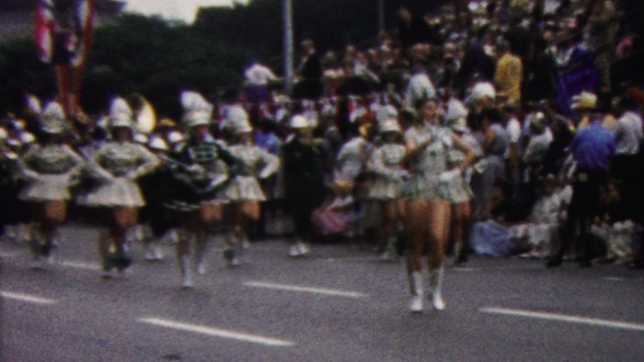 In 1961 California home-movie footage, white-uniformed marching performers and tall-hatted band members proceed down a street lined with spectators.