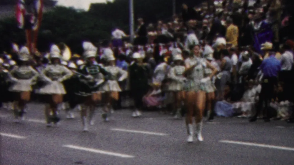 In 1961 California home-movie footage, white-uniformed marching performers and tall-hatted band members proceed down a street lined with spectators.