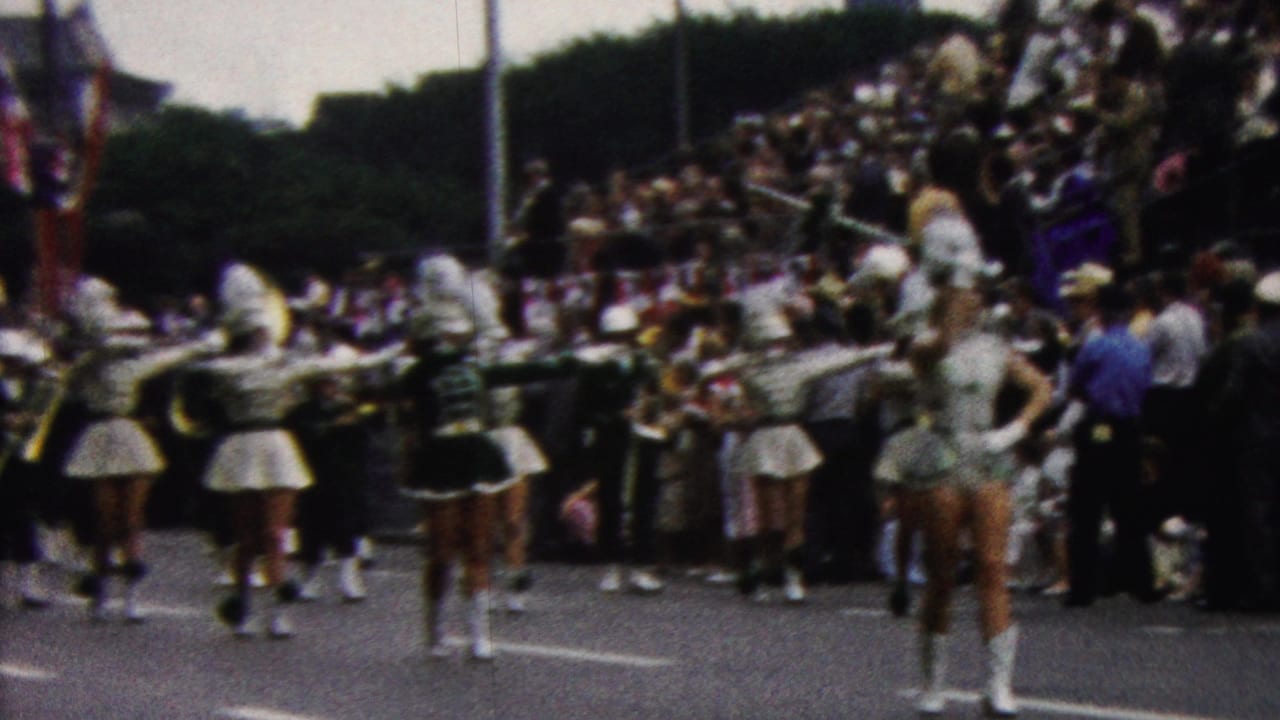 Band members and majorettes march past a crowded roadside viewing area.
