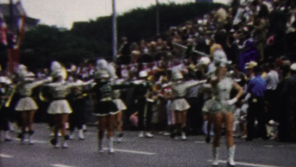 Band members with instruments and white-uniformed majorettes march in formation past spectators gathered along a California parade route in 1961.