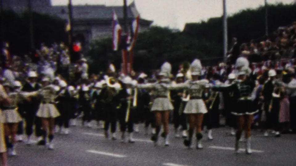 A marching band moves away from the camera as spectators, flags, and roadside decorations frame the 1961 California parade scene.