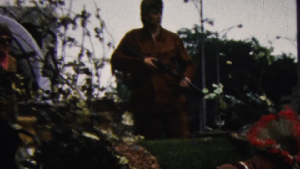 A man dressed in a Davy Crockett or frontier-style costume stands near parade decorations, with a feathered headdress visible in the foreground of the 1961 California home movie.